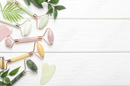 Face rollers, gua sha tools and green leaves on white wooden background, flat lay. Space for text photo