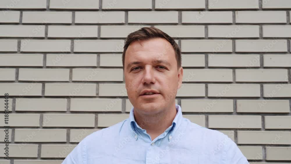 Adult caucasian man in blue shirt looking at camera and speaking against red brick wall background. Close-up shot of millennial guy. People and emotions concept. Adult caucasian man in blue shirt looking at camera and speaking against red brick wall background. Close-up shot of millennial guy. People and emotions concept.