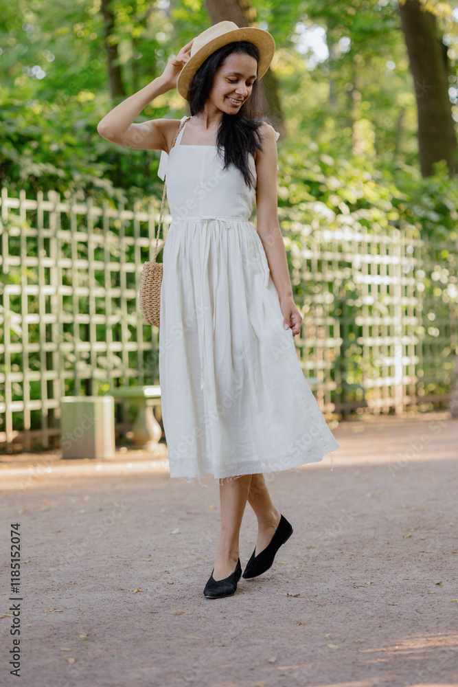 carefree African American woman in white dress and straw hat walks in park and poses against background of green hedge. daytime for relaxation. beautiful female fashion model, sunlight summer time carefree African American woman in white dress and straw hat walks in park and poses against background of green hedge. daytime for relaxation. beautiful female fashion model, sunlight summer time