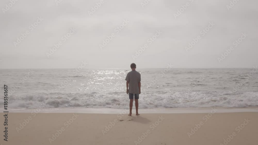 Teenager standing on sandy coastline, gazing at rolling waves during cloudy summer day, embodying peaceful seascape solitude. Spain, Galicia Teenager standing on sandy coastline, gazing at rolling waves during cloudy summer day, embodying peaceful seascape solitude. Spain, Galicia