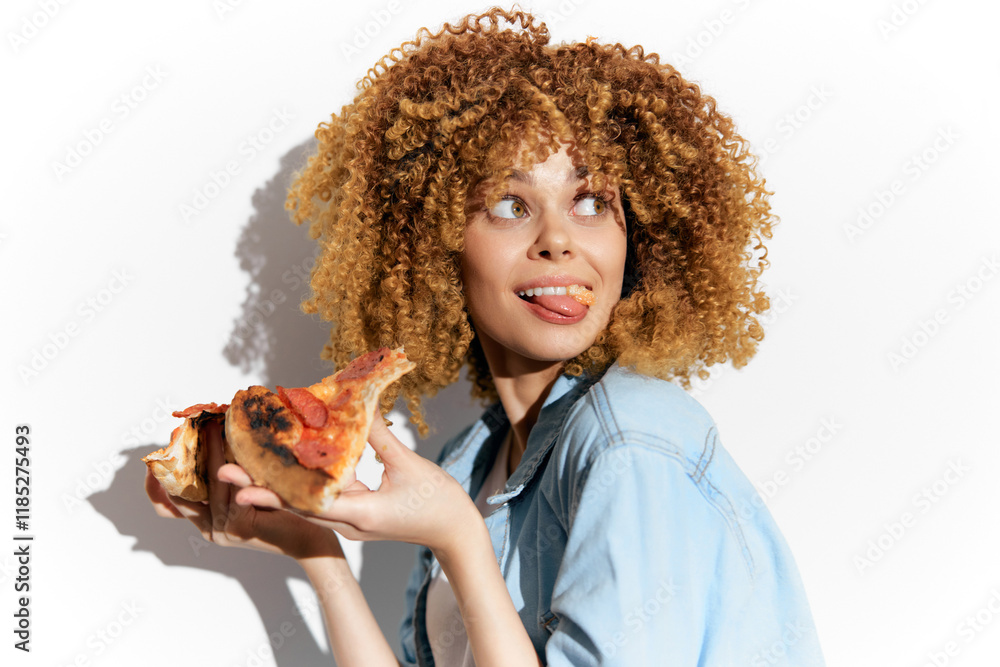 Playful young woman with curly hair enjoying a slice of pizza with a cheerful expression against a light backdrop, highlighting a fun food concept