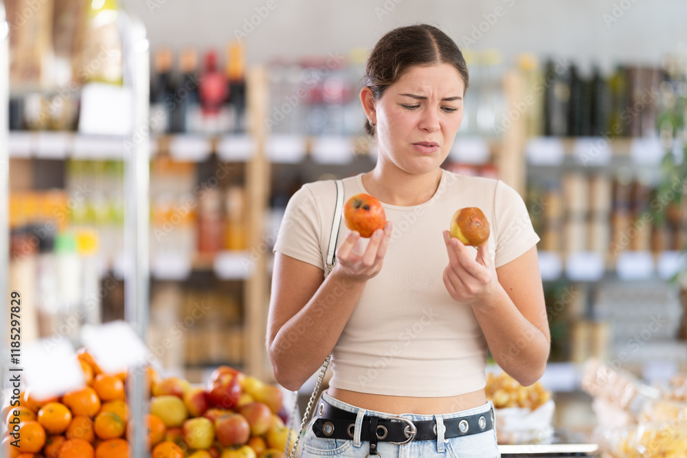 Fototapeta premium In store, frustrated girl holds rotten fruits in hands. She accidentally took spoiled fruits from showcase box, fastidiously examines unsuitable goods.