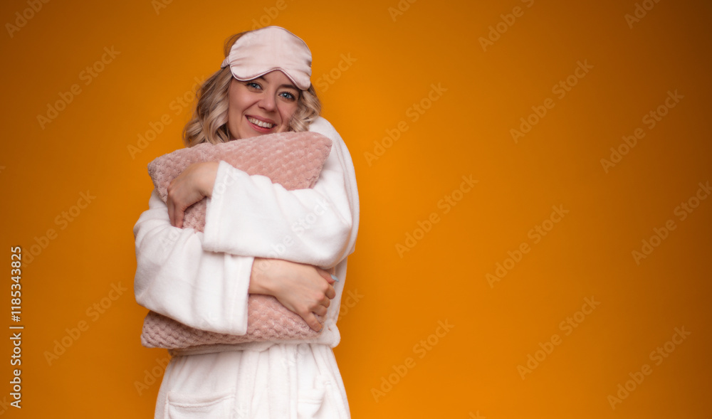 Fototapeta premium Woman in white robe and pink sleep mask, smiling and hugging soft pink pillow, set against a vibrant orange background. The image exudes warmth, comfort, and positivity, perfect for cozy home themes