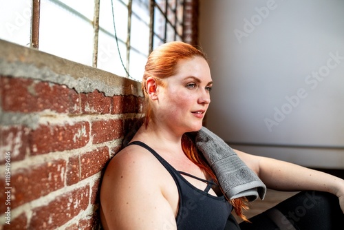 Woman resting after workout, sitting by brick wall. Fitness and relaxation. Red hair plus size woman in athletic build. Casual gym setting, towel on woman's shoulder. Post-exercise calm. photo