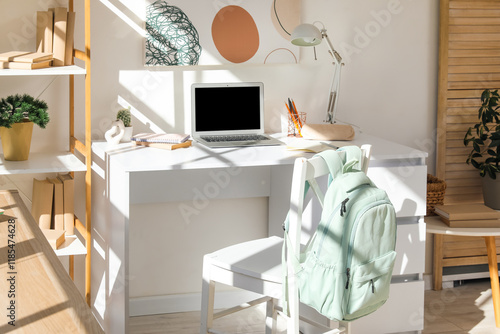 Modern school desk with stationery, backpack and laptop in room near white wall