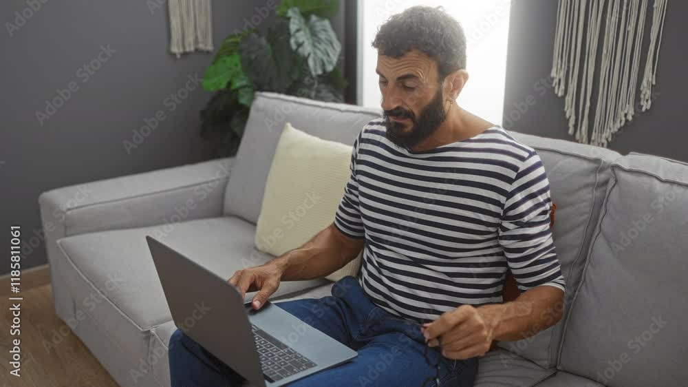 Young man sitting on a couch in a living room with a laptop, wearing a striped shirt, rubbing his eyes while holding his glasses, showing fatigue from working indoors at home Young man sitting on a couch in a living room with a laptop, wearing a striped shirt, rubbing his eyes while holding his glasses, showing fatigue from working indoors at home