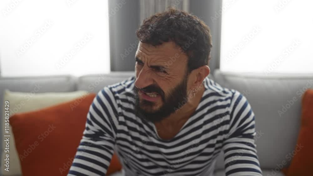 Frustrated man with a beard in a striped shirt sitting on a couch in a living room Frustrated man with a beard in a striped shirt sitting on a couch in a living room