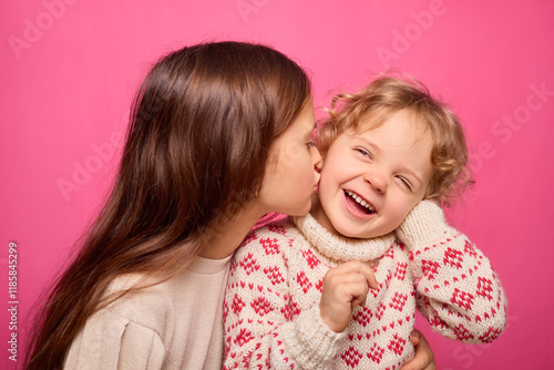 A joyful young girl laughs while her older sister kisses her cheek. They are in front of a pink background, showcasing a warm and loving sibling bond during playtime photo