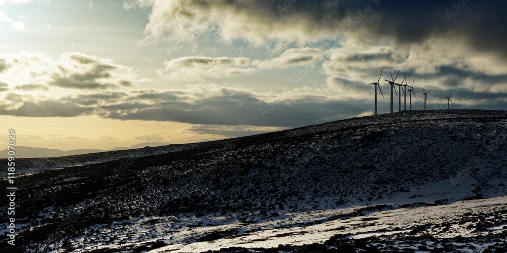 Group of wind turbines on a snowy hill under a cloudy sky, with the sun shining through the clouds Group of wind turbines on a snowy hill under a cloudy sky, with the sun shining through the clouds