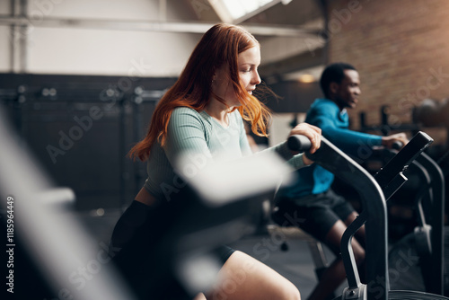 Diverse friends working out during a gym stationary bike class. The woman's red hair is moving in the wind from the bicycle. Her friend is focused on the bike.