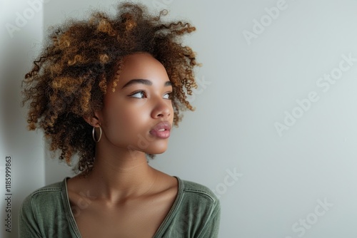 Young woman with curly hair gazes thoughtfully near a white wall in natural light photo