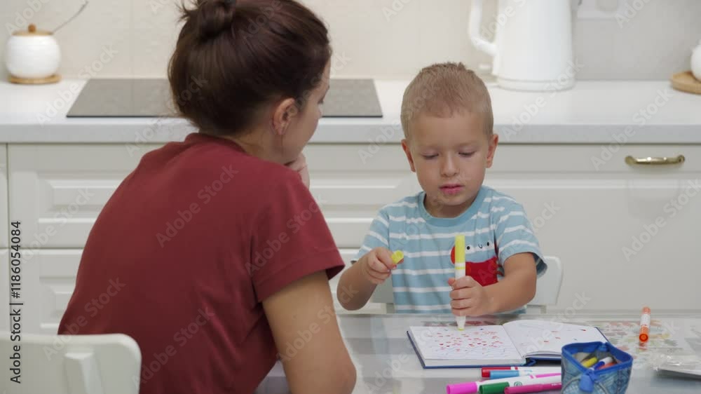 Mom and son coloring on kitchen table. Mother is watching her son color in coloring book in kitchen. Child and mother have fun and spend time together. Mom and son coloring on kitchen table. Mother is watching her son color in coloring book in kitchen. Child and mother have fun and spend time together.
