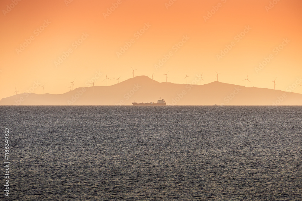 Cargo ship with wind turbines on a mountain in foreground. Concept image for transportation and green energy industry. Sunset landscape photo over the sea.