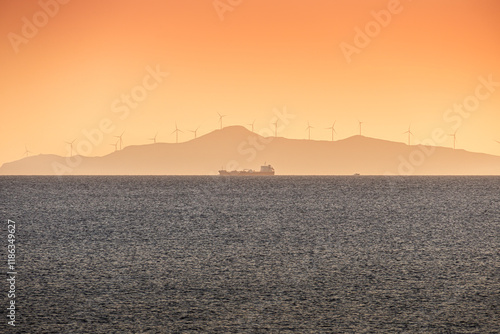 Obraz na płótnie Cargo ship with wind turbines on a mountain in foreground. Concept image for transportation and green energy industry. Sunset landscape photo over the sea. w salonie