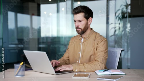 Frustrated businessman having difficulty making transaction using laptop while sitting at desk at workplace in a modern business office. Confused male worker having problems working online on computer