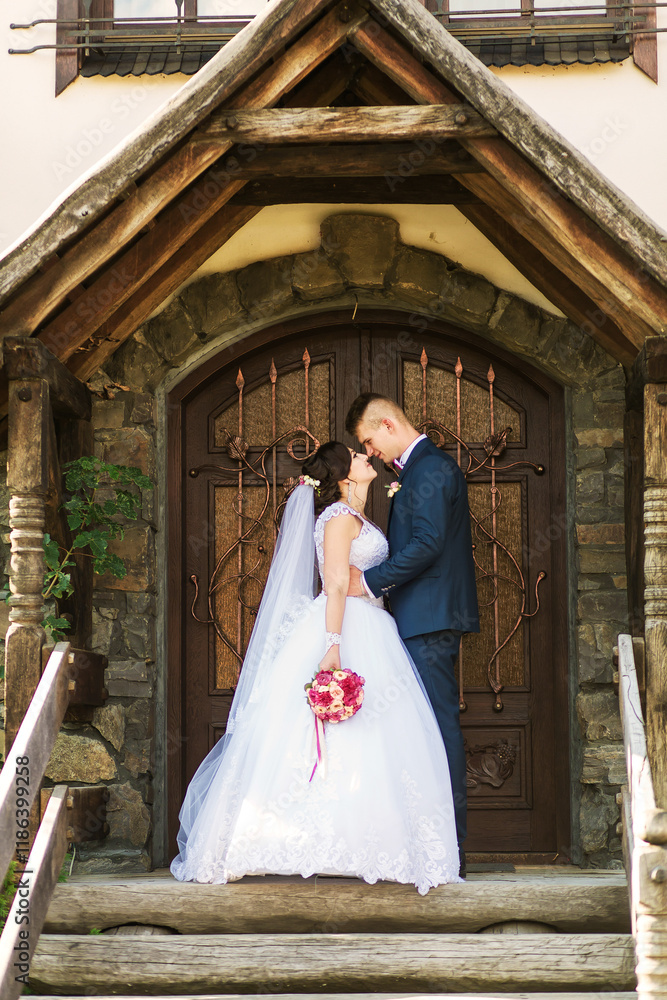 Fototapeta premium A bride and groom are standing in front of a wooden door, with the bride holding a bouquet. Scene is romantic and intimate, as the couple is embracing each other