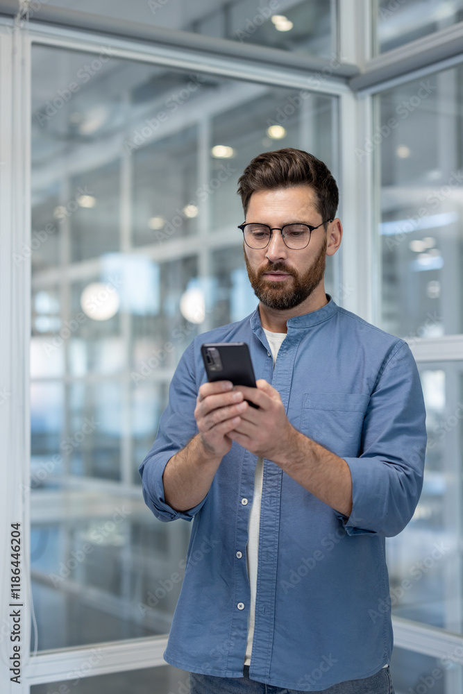 Serious confident man with phone in hands, businessman standing near window inside office at workplace, using app on smartphone, typing text message. Serious confident man with phone in hands, businessman standing near window inside office at workplace, using app on smartphone, typing text message.