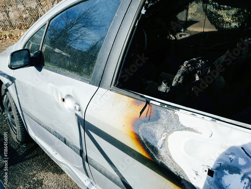 Close-up of a silver car with visible fire damage, including charred and burnt areas on the door and window, parked outdoors on a sunny day.