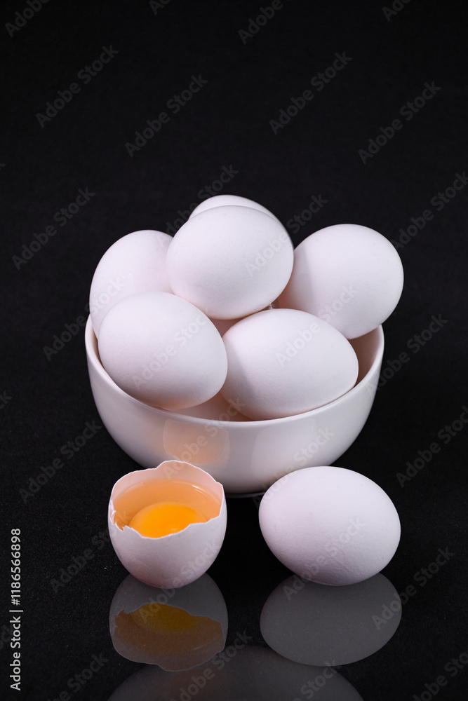 The image shows a white bowl filled with chicken eggs against a black background. Next to the misska there is a whole and a broken egg. The image shows a white bowl filled with chicken eggs against a black background. Next to the misska there is a whole and a broken egg.