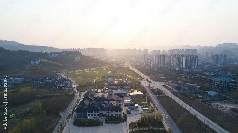 Fototapeta premium Sunrise Over Cityscape: An aerial view captures the sun rising over a sprawling cityscape nestled between rolling hills. The hazy morning light bathes the scene in a golden glow.