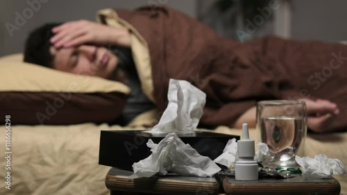 A man is lying very sick in bed, looking miserably ill. Medicines, water and napkins are in the foreground, symbolizing the concept of illness photo