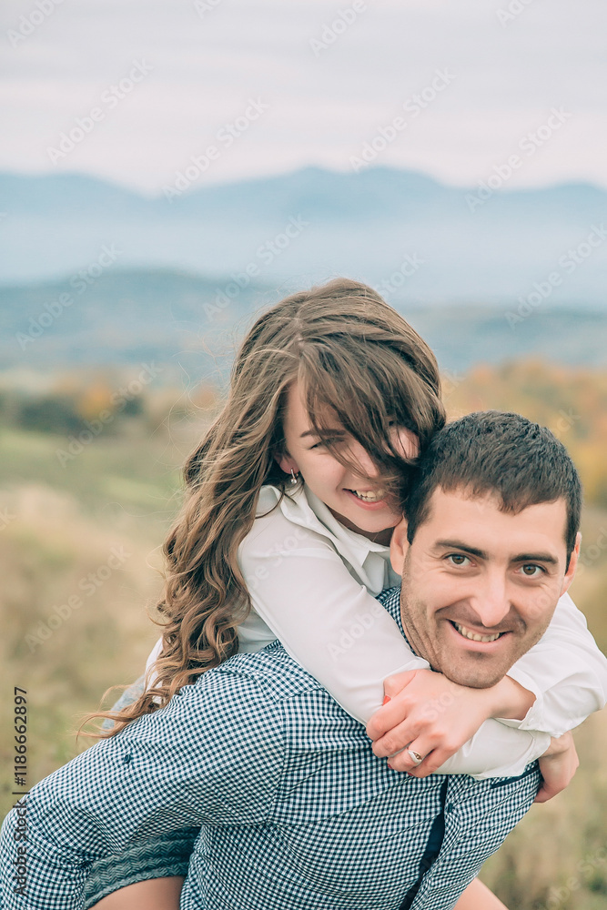 A man and a woman are standing on a grassy hillside, with the man carrying the woman on his back. The man is smiling and the woman is laughing, creating a joyful and lighthearted atmosphere A man and a woman are standing on a grassy hillside, with the man carrying the woman on his back. The man is smiling and the woman is laughing, creating a joyful and lighthearted atmosphere