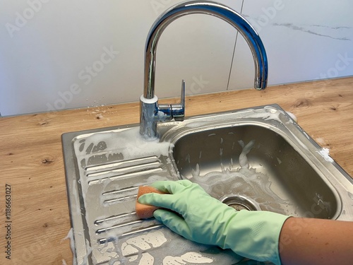 A hand in a rubber glove washes the kitchen sink with a sponge, covered in foam from detergent. Ideal for cleaning, home maintenance, and detergent-related themes. Keep your space clean and fresh.
