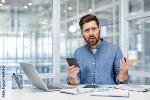 Upset and disappointed man with phone in hands. Office worker at workplace looking anxiously at camera, working with laptop inside office at workplace. photo