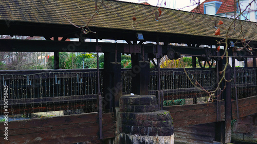 A river flows through which an old wooden bridge with a roof passes. The bridge is located in Europe and stands on wooden and concrete structures. On the edges of the river there are old stone walls p photo