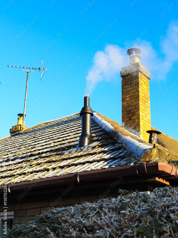 Naklejka premium A newly fitted flue pipe from a burning heating boiler, expelling fumes through a frosty domestic house roof, next to a chimney stack and TV aerial, on a cold winter morning, against a blue sky.