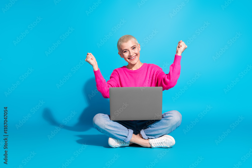 Young woman wearing a vibrant pink sweater and light blue jeans happily uses a laptop while sitting cross-legged Young woman wearing a vibrant pink sweater and light blue jeans happily uses a laptop while sitting cross-legged