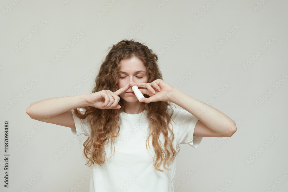 Fototapeta premium A young woman with curly hair in a white T-shirt uses a nasal spray, looking anxious and confused. Concept of health care, allergies, self-treatment, and battling cold or respiratory symptoms.