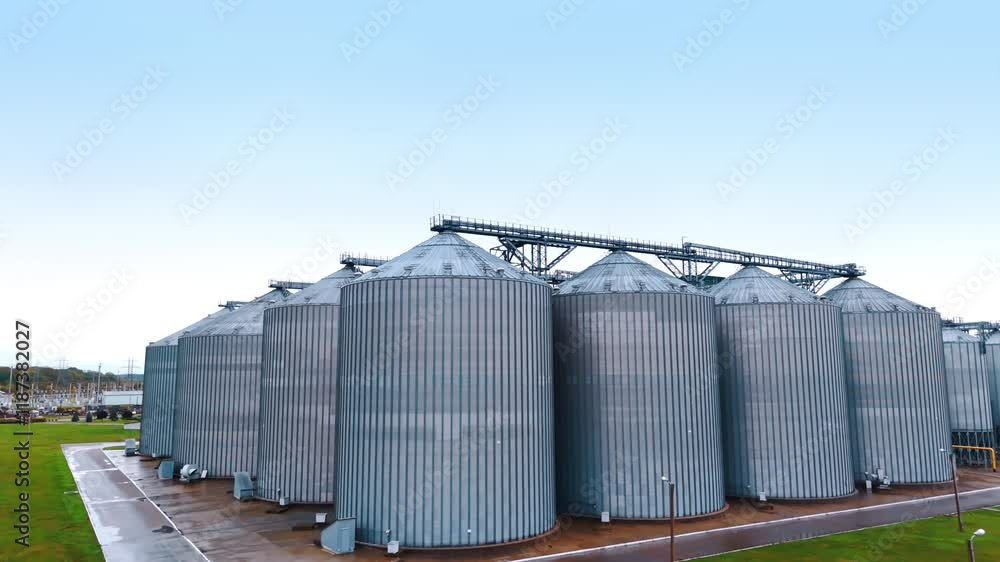 Huge silver silos at agro manufacturing plant for processing, drying, cleaning and storage of agricultural products, flour, cereals and grain. Territory of the modern plant in the rural area. Huge silver silos at agro manufacturing plant for processing, drying, cleaning and storage of agricultural products, flour, cereals and grain. Territory of the modern plant in the rural area.
