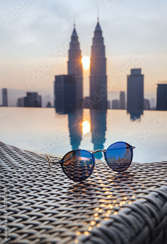 Photography Fancy sunglasses forgotten on sanbed next to infinity pool with two skyscrapers tower twins reflected on water surface during evening sunset time