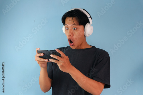 Portrait of Asian young man in black t-shirt using wireless headphone playing games on his mobile phone by tilting the screen. Wow face expression photo