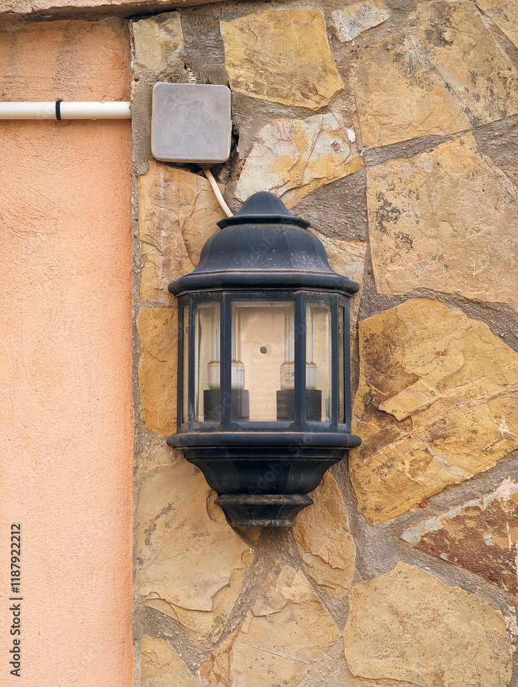 Close-up of black metal unlit rustic wall lantern against natural stone textured facade by day, Mediterranean outdoor decor style. Close-up of black metal unlit rustic wall lantern against natural stone textured facade by day, Mediterranean outdoor decor style.