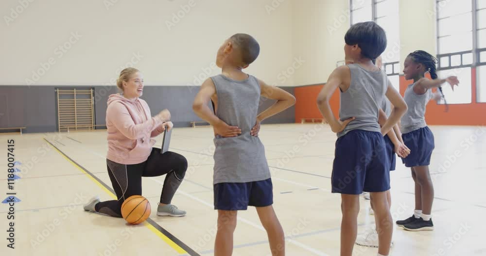 In school gym, coach kneeling with basketball, instructing students during practice In school gym, coach kneeling with basketball, instructing students during practice