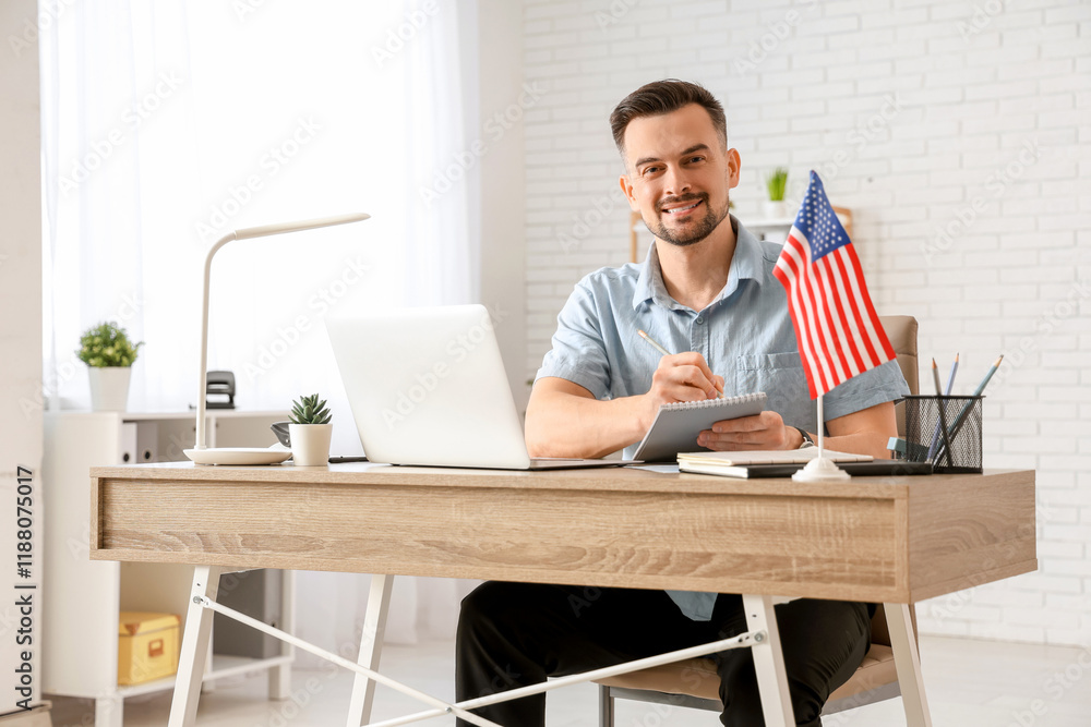 Handsome young happy man with laptop and flag of USA writing in notebook at office Handsome young happy man with laptop and flag of USA writing in notebook at office