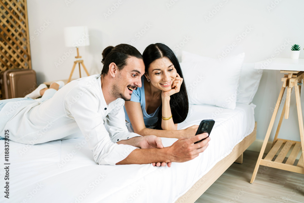 A Caucasian couple enjoys quality time together, sharing smiles while looking at a smartphone in a cozy bedroom setting. Couple spend quality time in a relationship. A Caucasian couple enjoys quality time together, sharing smiles while looking at a smartphone in a cozy bedroom setting. Couple spend quality time in a relationship.