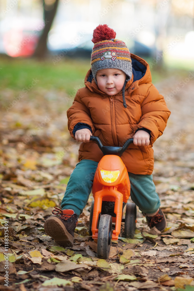 A small child wearing a warm orange jacket and a colorful hat rides a bright orange tricycle on a path covered with fallen leaves. The cheerful atmosphere reflects a sunny autumn day. A small child wearing a warm orange jacket and a colorful hat rides a bright orange tricycle on a path covered with fallen leaves. The cheerful atmosphere reflects a sunny autumn day.