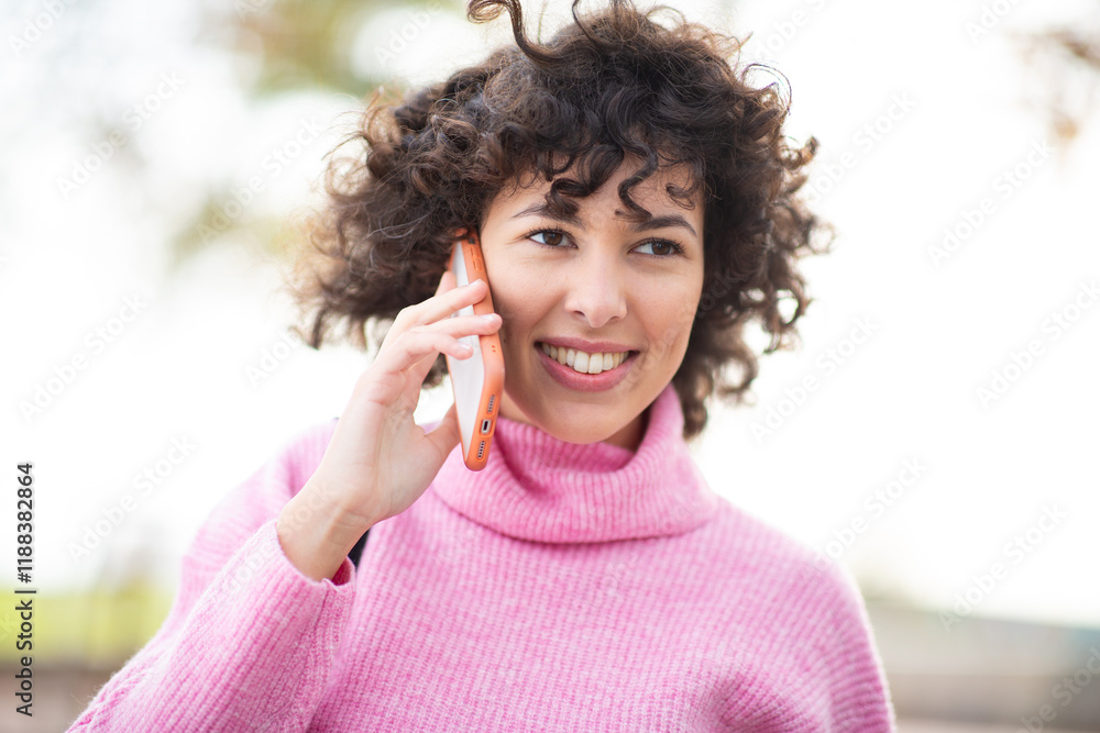 Young woman with curly hair and pink turtleneck sweater talking on her phone outdoors Young woman with curly hair and pink turtleneck sweater talking on her phone outdoors
