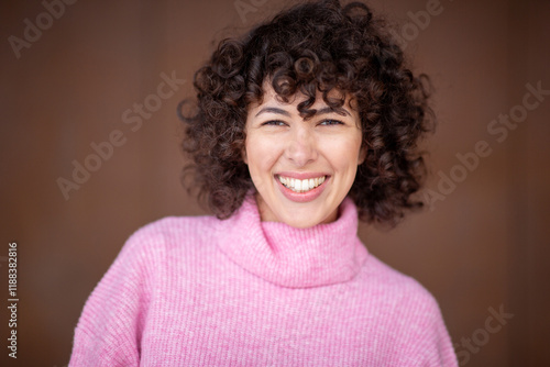 Close-up of a cheerful woman with curly hair wearing a pink turtleneck sweater and smiling directly at the camera, isolated on a blurred brown background