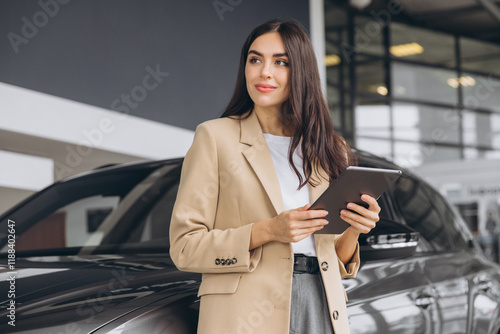 Smiling car seller, businesswoman in beige suit standing in car salon and using tablet for choosing right car. photo