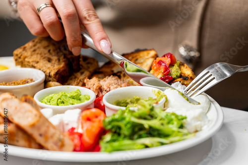 Wallpaper Mural A close-up of a hand holding a fork and knife, cutting into a colorful plate of food. The dish features vegetables, sauces, and slices of bread, showcasing a delicious meal. Torontodigital.ca