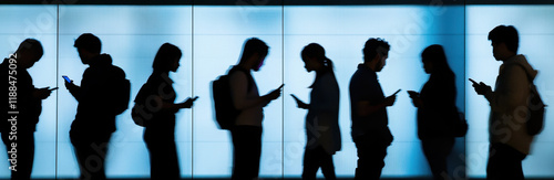 A group of people stands in line, illuminated by a bright backdrop, each person focused on their smartphone.