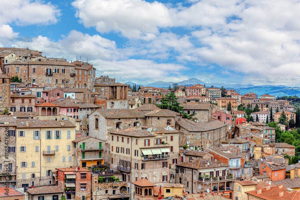 Elevated view reveals the tiled roofs of medieval buildings in Perugia, surrounded by lush greenery and distant mountains, embodying the charm of Umbria.