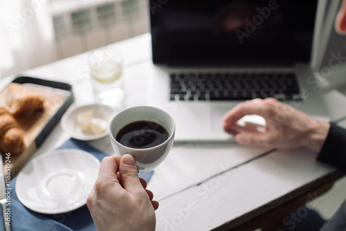 Close-up of a man drinking coffee and eating croissants while using a laptop computer