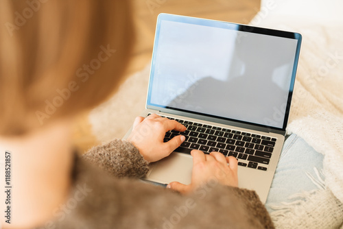 View from behind of a young woman sitting on a comfortable sofa in her living room using laptop computer with blank white screen