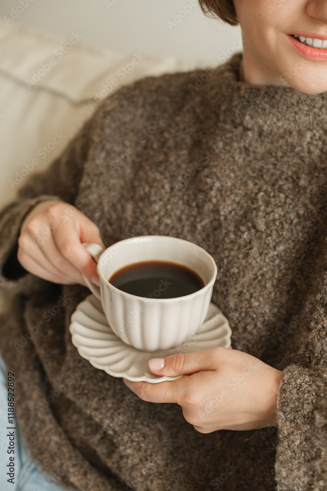 Close-up of a young woman in a woolly jumper sitting on her sofa and holding a cup of black coffee Close-up of a young woman in a woolly jumper sitting on her sofa and holding a cup of black coffee