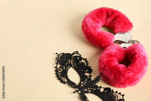 Pink fluffy handcuffs and lace mask on pale yellow background, flat lay. Space for text photo